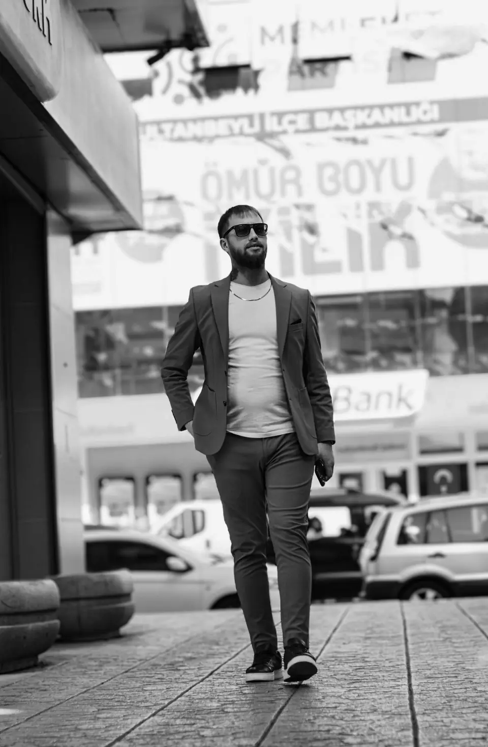 A bearded man in sunglasses and a blazer walks toward the camera along a paved city sidewalk.