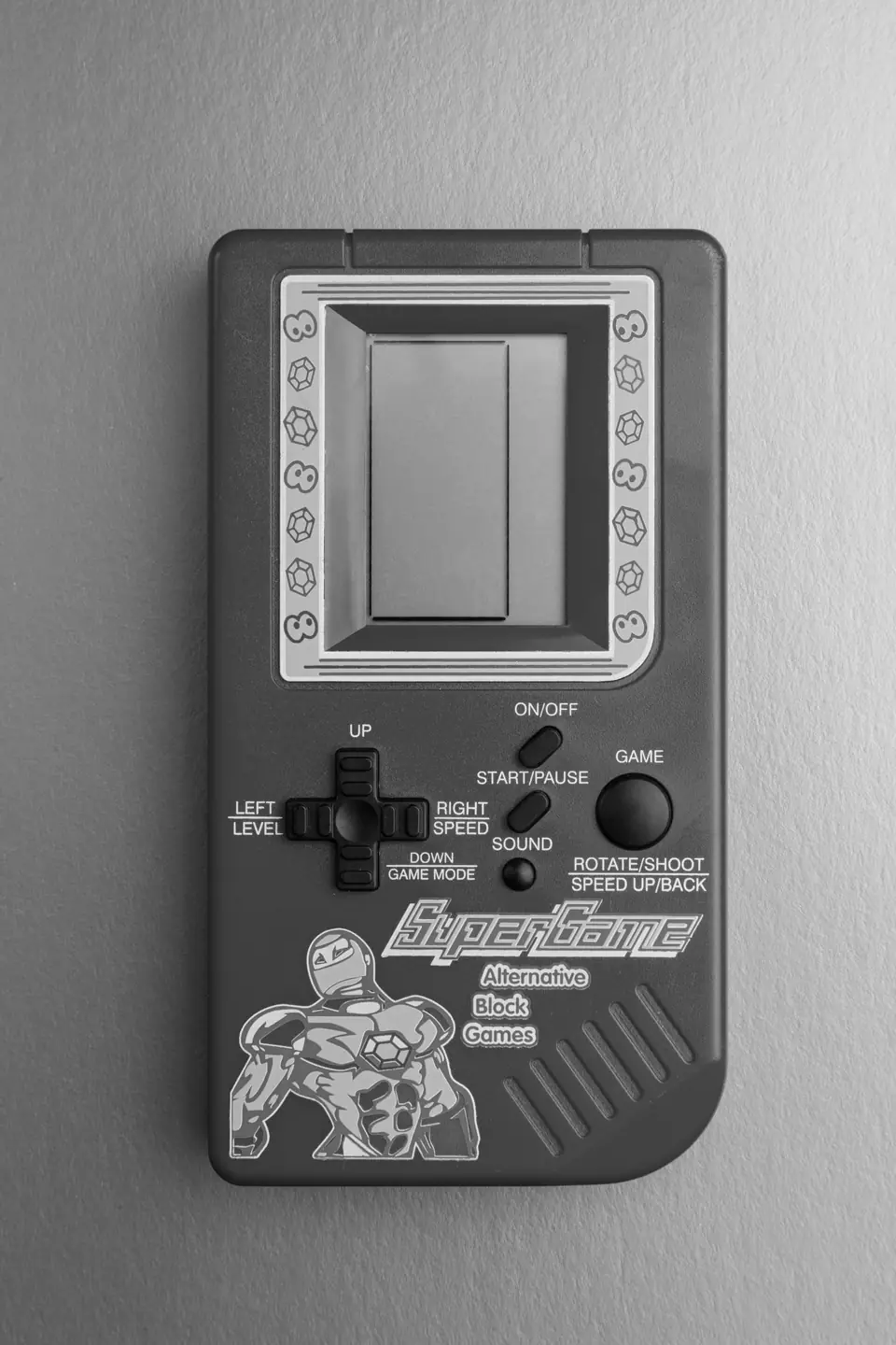 A gray handheld brick game device with a small screen, direction pad and cartoon hero decal lies flat.