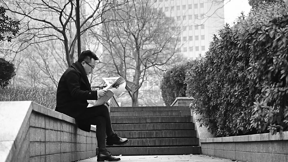 A man in a dark coat and glasses sits on a low stone wall reading a newspaper beside outdoor steps.