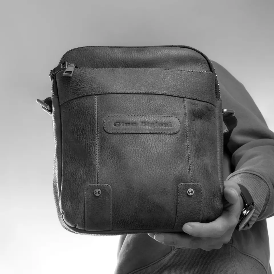 A person holds up a textured leather crossbody bag with a zippered front pocket and an embossed label.