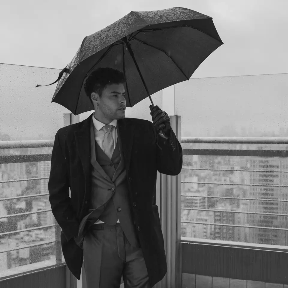 A man in a suit and overcoat holds an open umbrella on a rooftop terrace above a city.