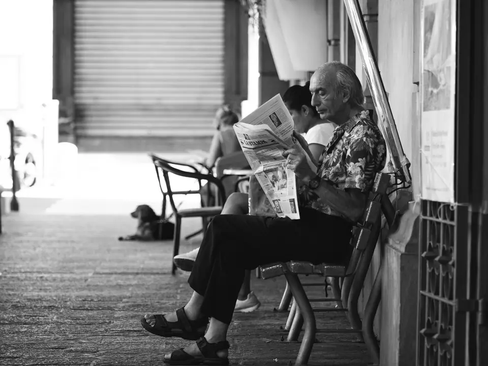 An older man in a patterned shirt and sandals reads a newspaper on a café bench beside a sleeping dog.