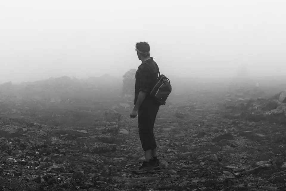A man with a small backpack stands on a stony plain that fades into fog.