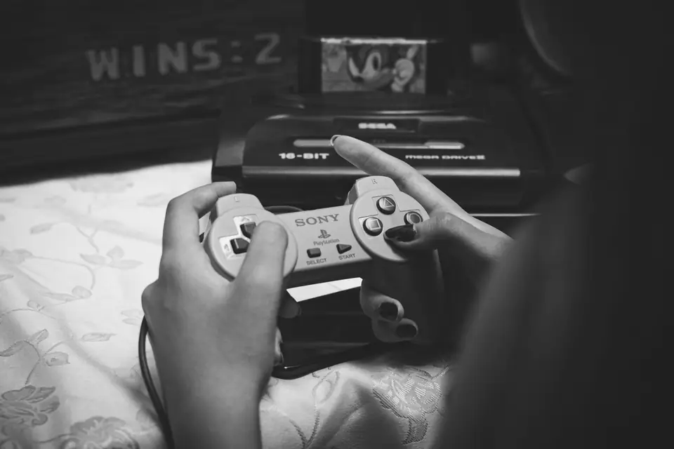 A person on a bed holds a game controller in front of a black 16-bit console.