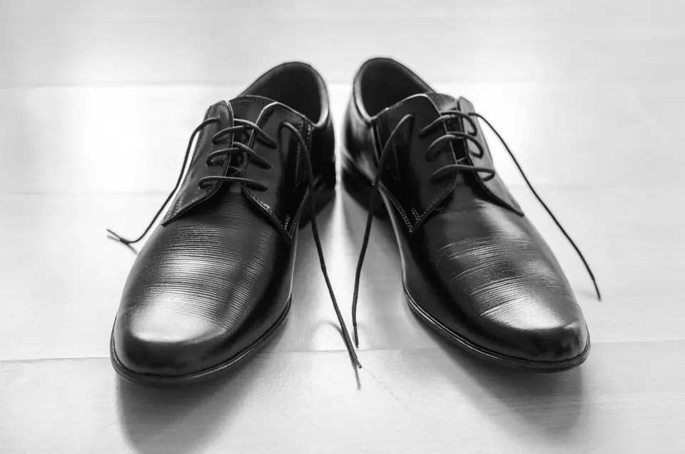 A pair of shiny black lace-up oxford shoes sits on a light wooden floor with untied laces.