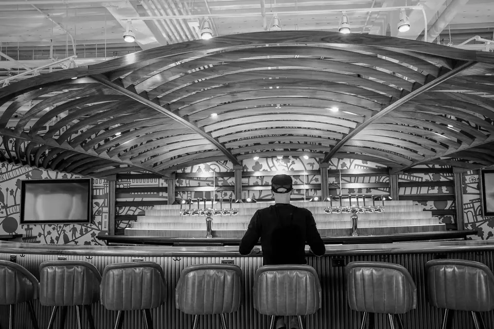 A man in a cap sits alone at a long bar with empty stools under a curved wooden ceiling.