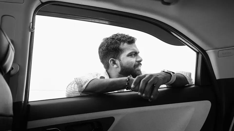 A bearded man rests his arms on an open car window and gazes off to one side.