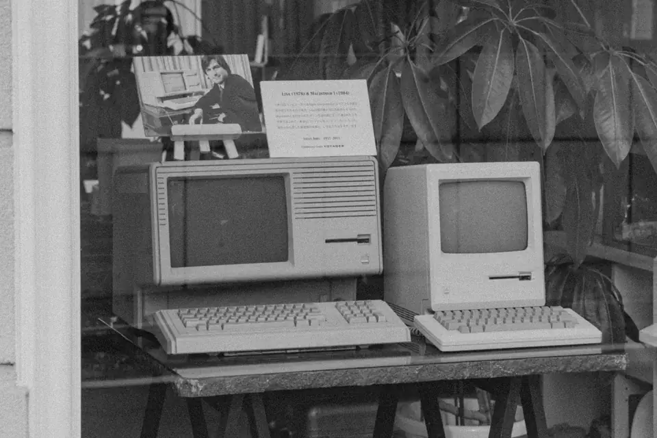 Two vintage desktop computers with keyboards sit on a table behind a window with a placard.
