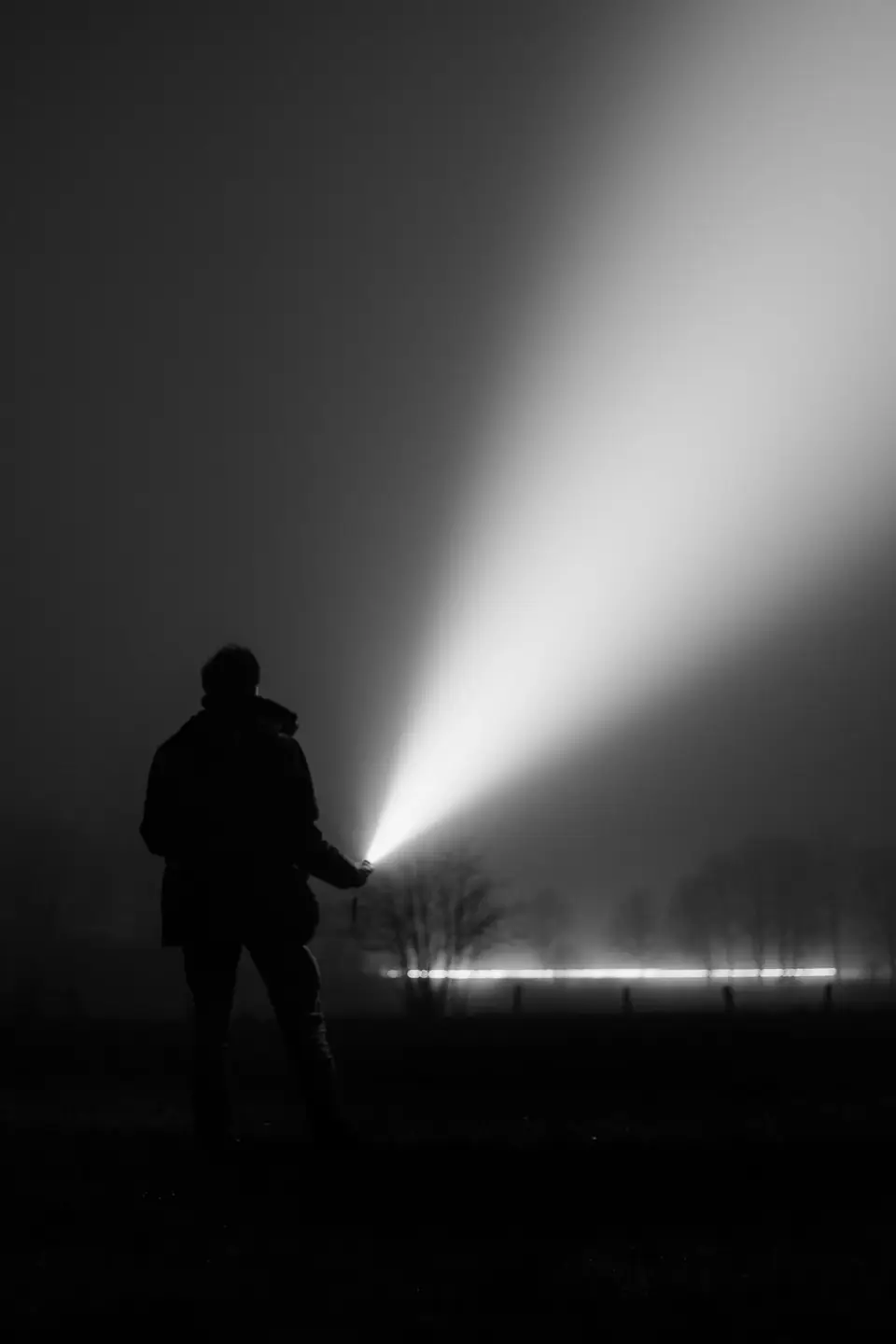 A silhouetted person stands in a foggy field at night holding a flashlight that casts a broad beam.