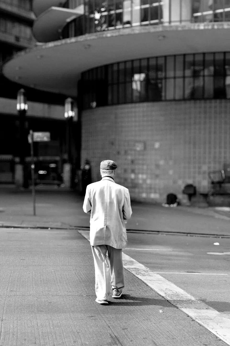 An elderly man in a light suit and flat cap walks away across a wide, empty city plaza.