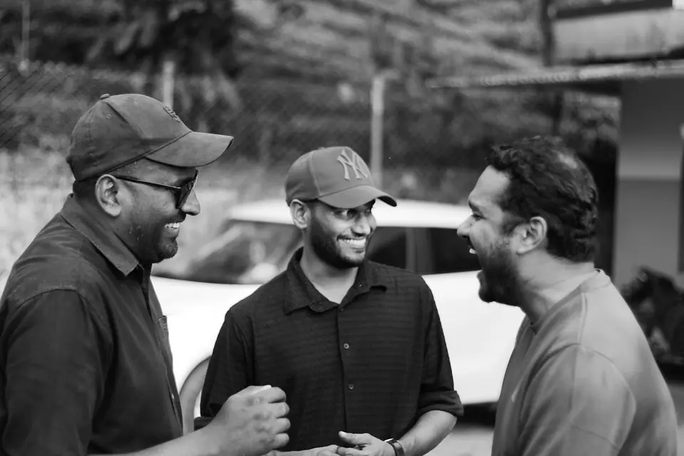 Three men in caps and casual shirts talk and laugh together outdoors near a parked white car.