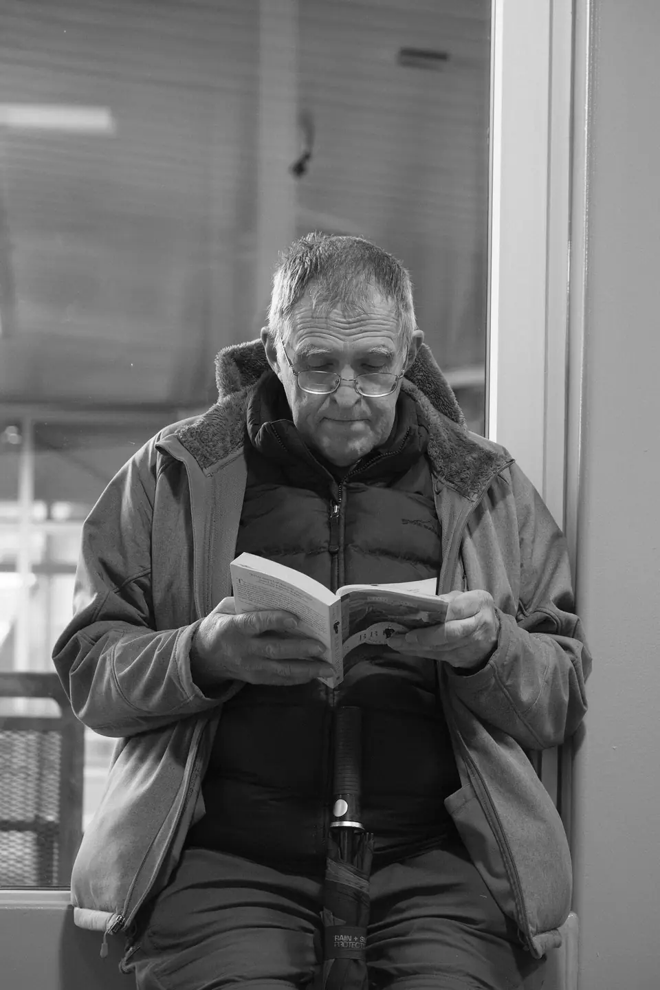 An older man in glasses and a padded jacket sits by a window reading a paperback book.