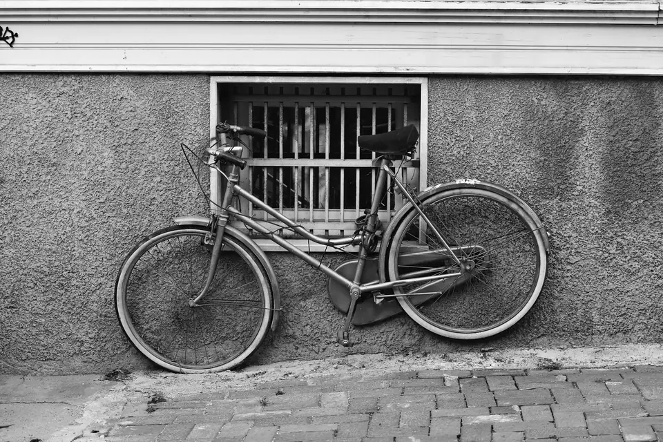 A weathered bicycle leans against a textured wall beneath a small barred window on a brick sidewalk.