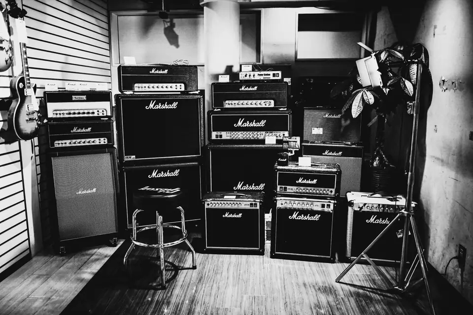 Stacked guitar amplifiers and speaker cabinets fill a room with a stool and a guitar.