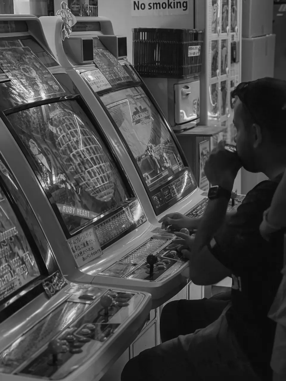 A man sits at an arcade cabinet in a row of machines beneath a “No smoking” sign.