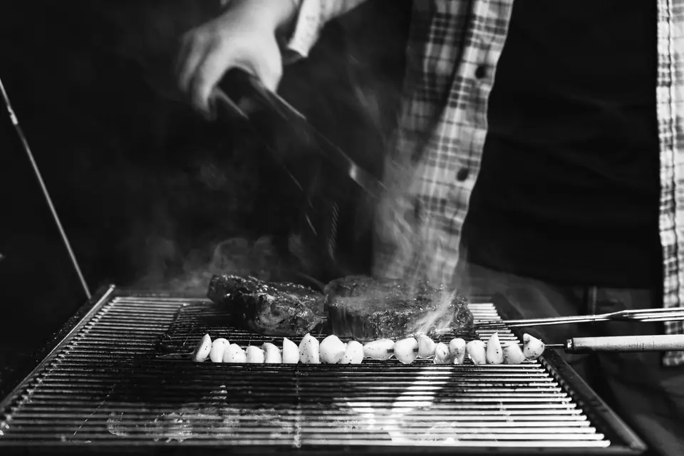 A person turns steaks with tongs on a smoking grill beside a row of vegetable skewers.