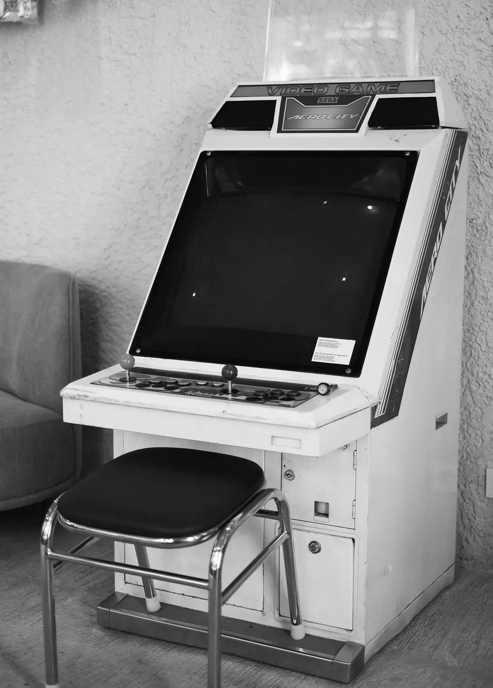 A white sit-down arcade cabinet with a dark screen stands beside a padded stool.