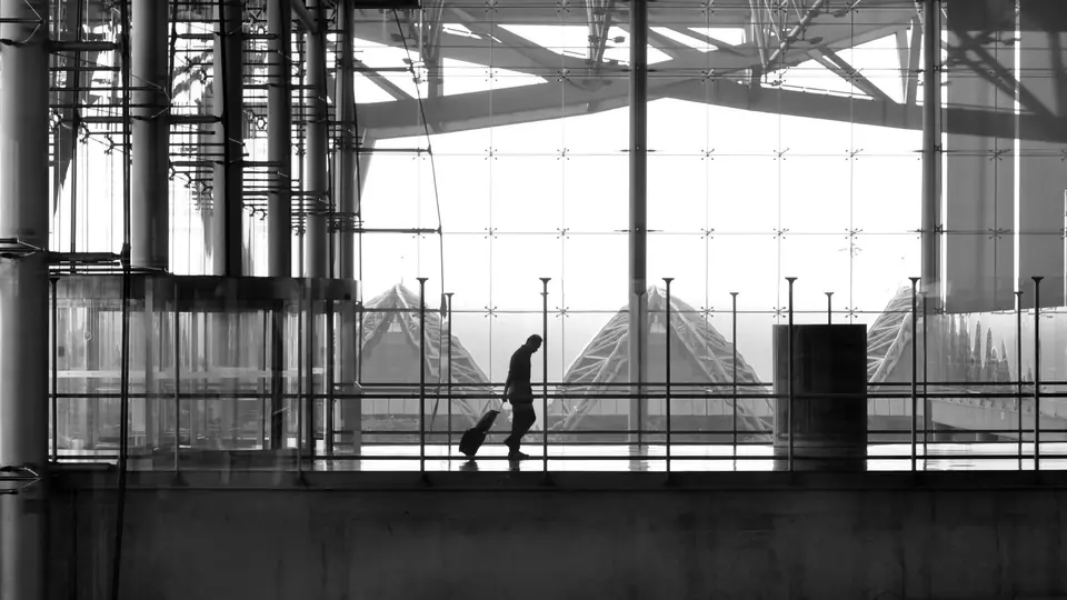 A silhouetted person pulls a small rolling suitcase along a walkway inside a large glass airport terminal.