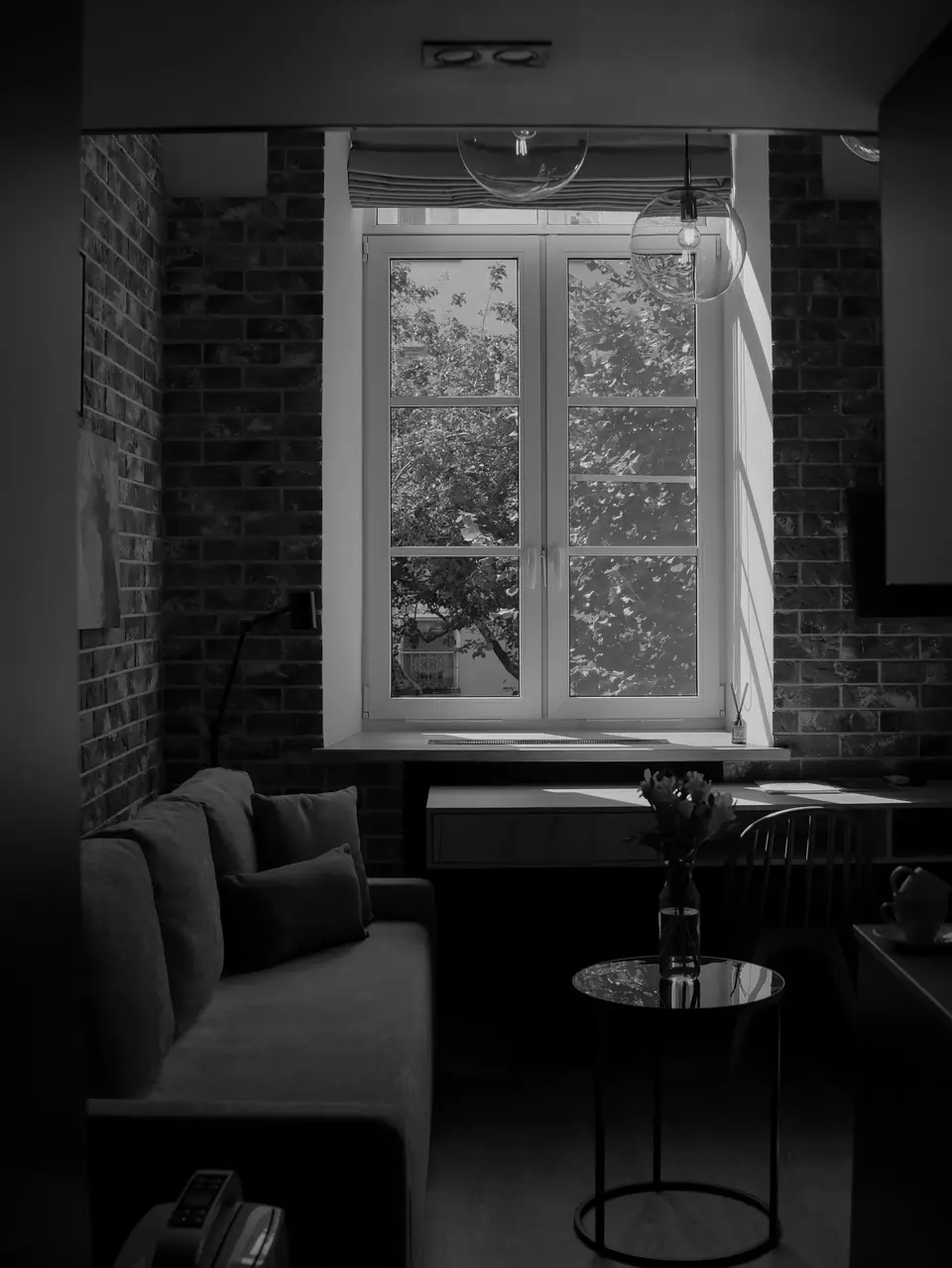 A small room with exposed brick has a sofa, a round side table and a desk beneath a tall window.
