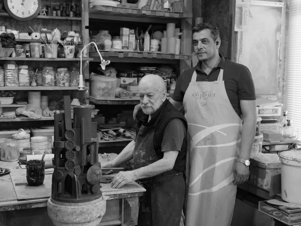 An elderly man in a vest and a younger man in an apron stand at a workbench in a cluttered workshop.