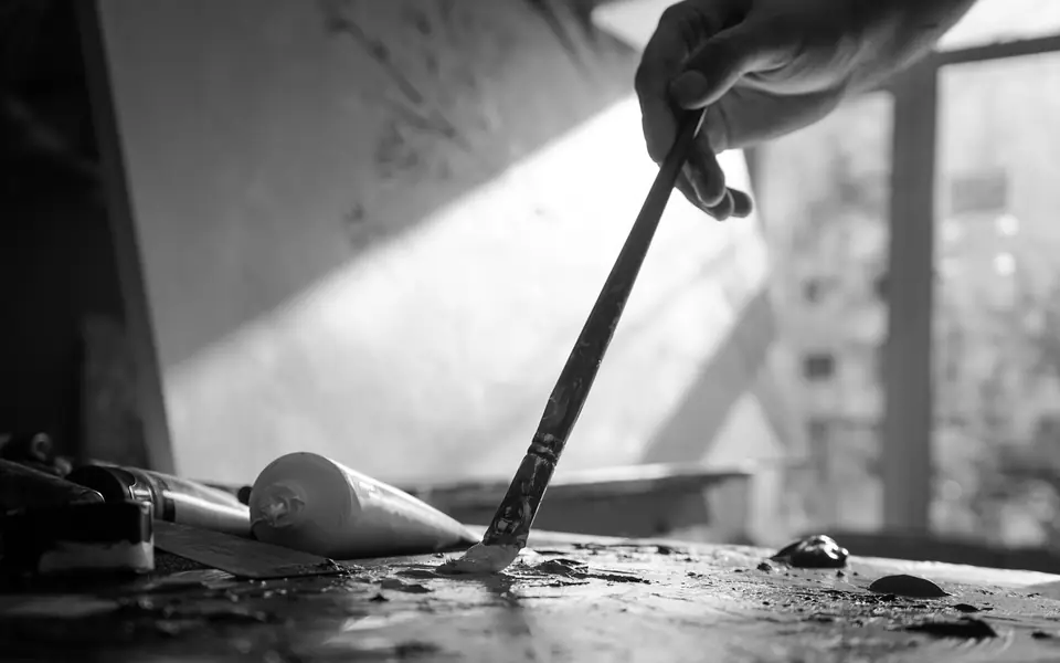 A hand dips a long paintbrush into paint on a cluttered studio table near a window.