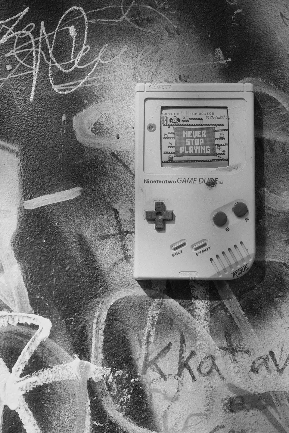 A street-art painting of a gray handheld console reading “Never Stop Playing” appears on a graffiti-covered wall.