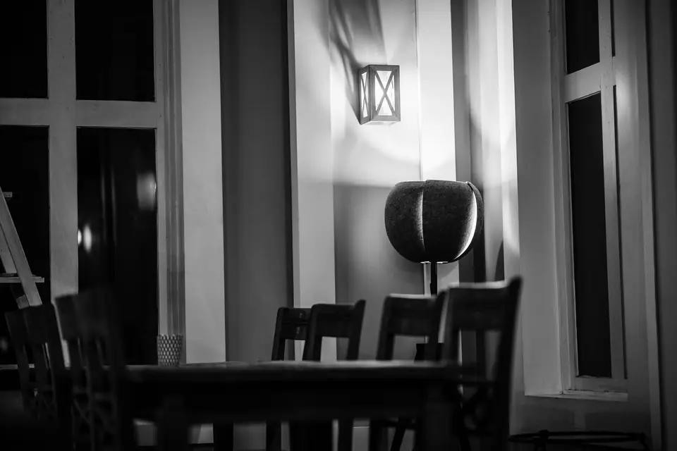 Wooden chairs stand around an empty table in a dim room lit by a single wall lamp.
