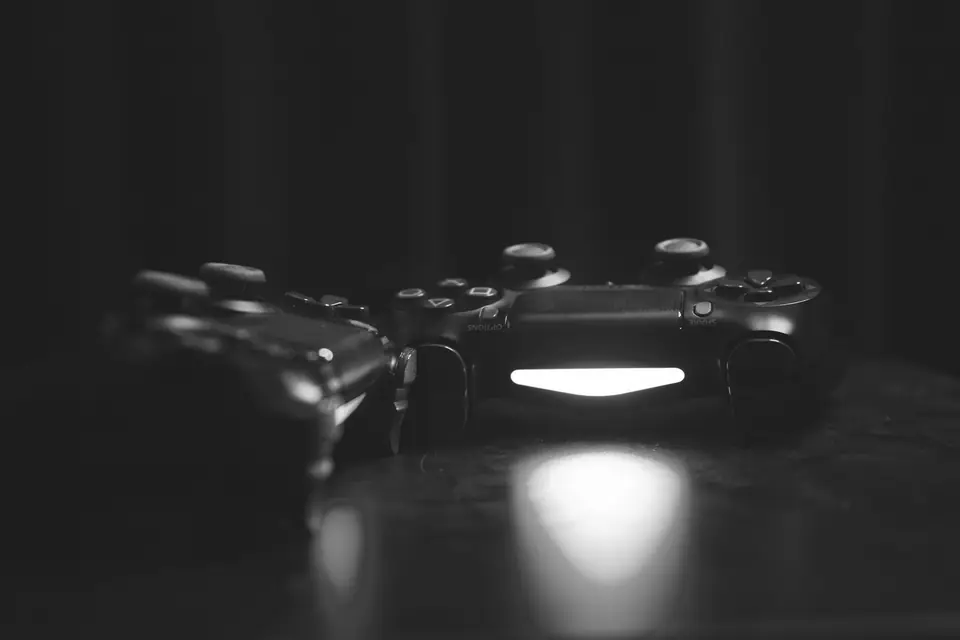 Two game controllers rest side by side on a dark surface, one with a glowing light bar.