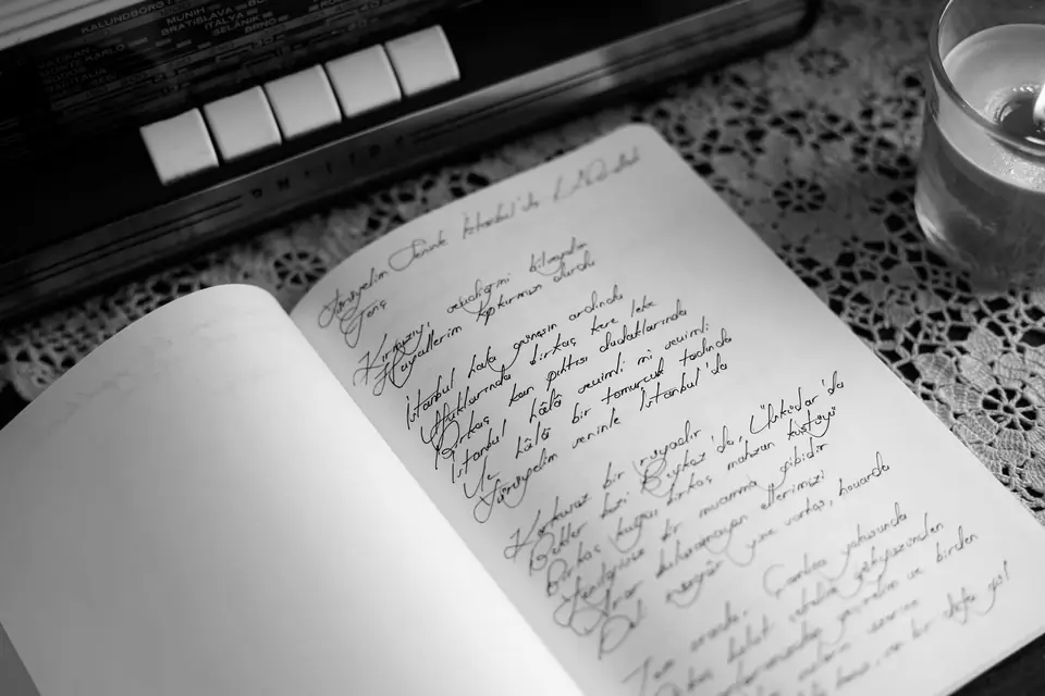 An open notebook filled with handwritten script lies on a lace tablecloth beside a glass.