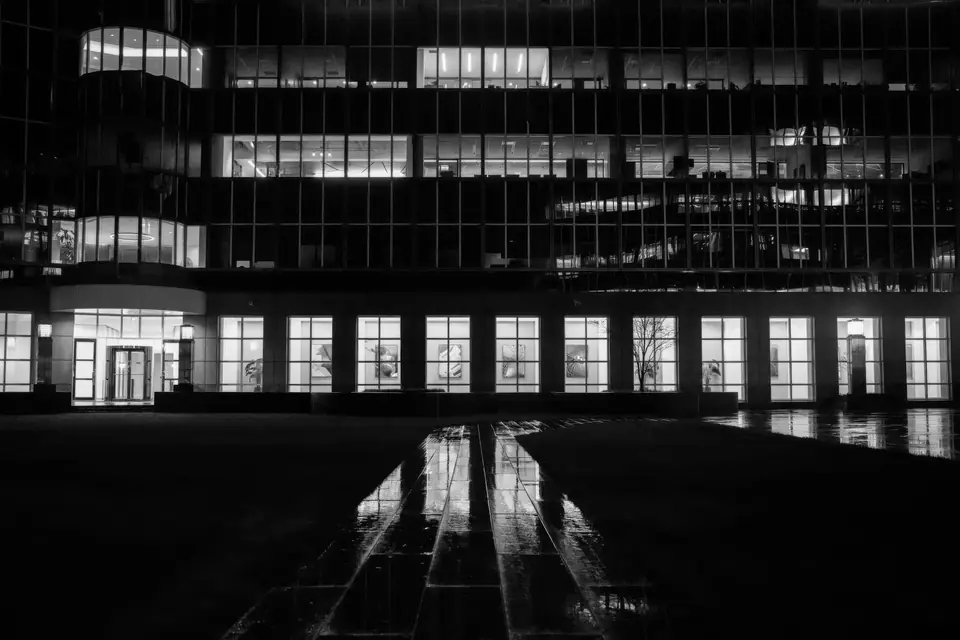 A modern office building at night with lit windows reflected on a wet walkway.
