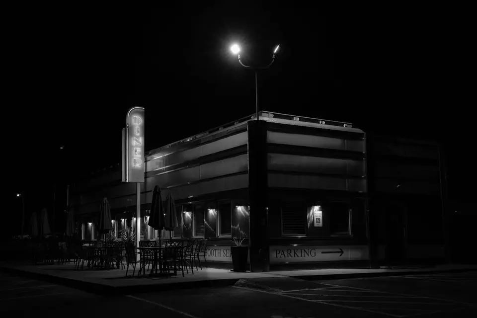 A diner with a vertical sign sits at the corner of a dark parking lot under a streetlamp.