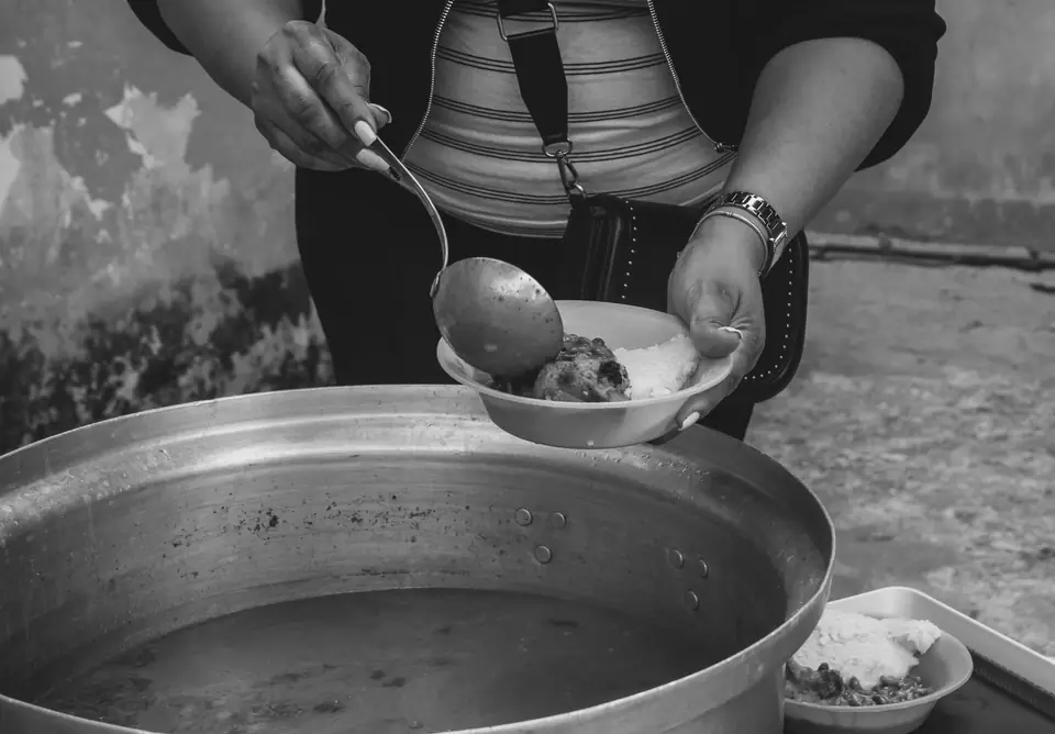 A person ladles stew from a large metal pot into a bowl held in their other hand.