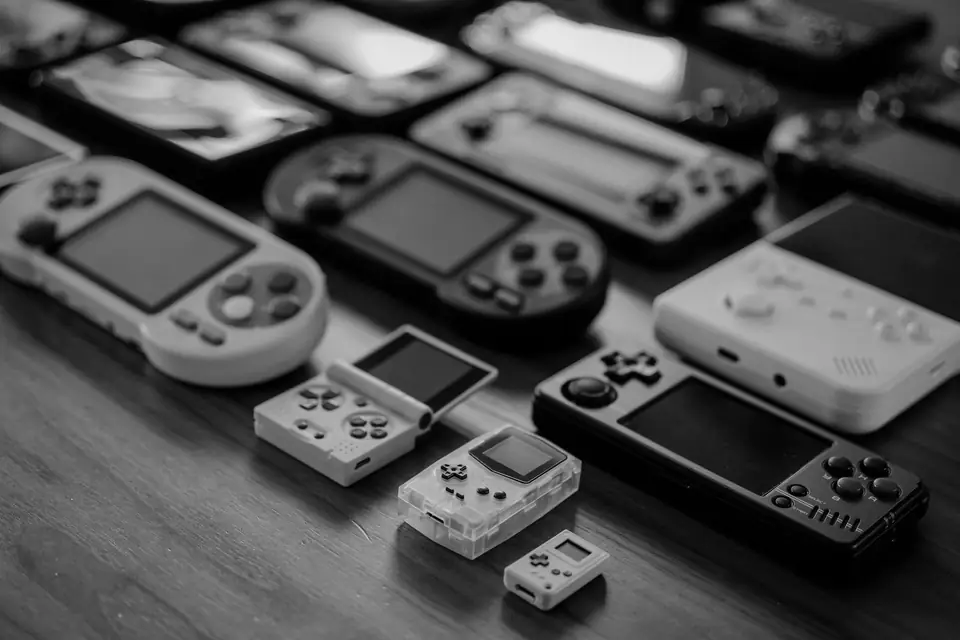 Rows of handheld game devices with small screens and buttons lie on a wooden table.
