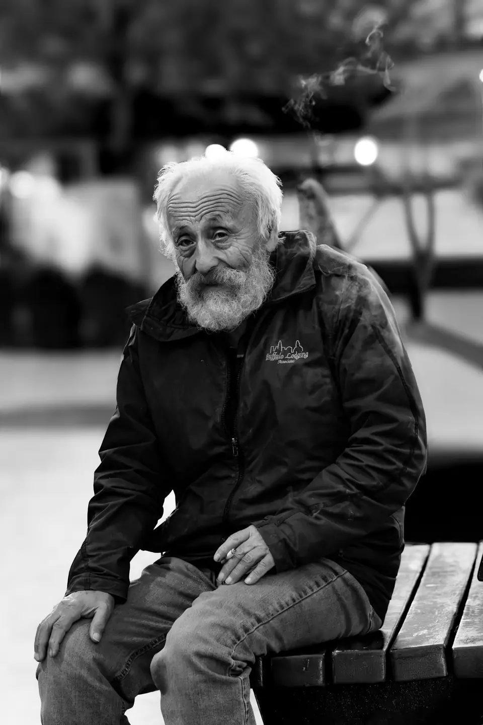 An elderly man with white hair and a beard sits on a wooden bench wearing a dark jacket.