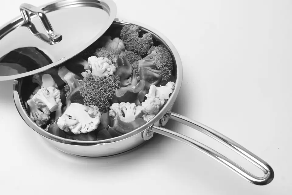 A stainless steel sauté pan filled with broccoli and cauliflower florets, its lid tilted open.