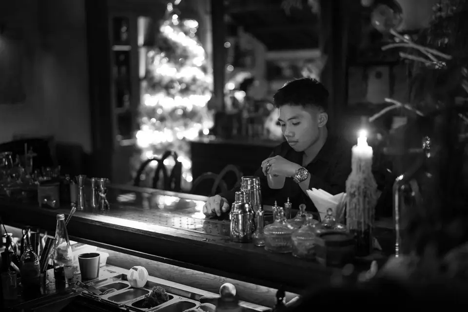 A young man sits alone at a bar lit by a candle, with a lit Christmas tree behind him.