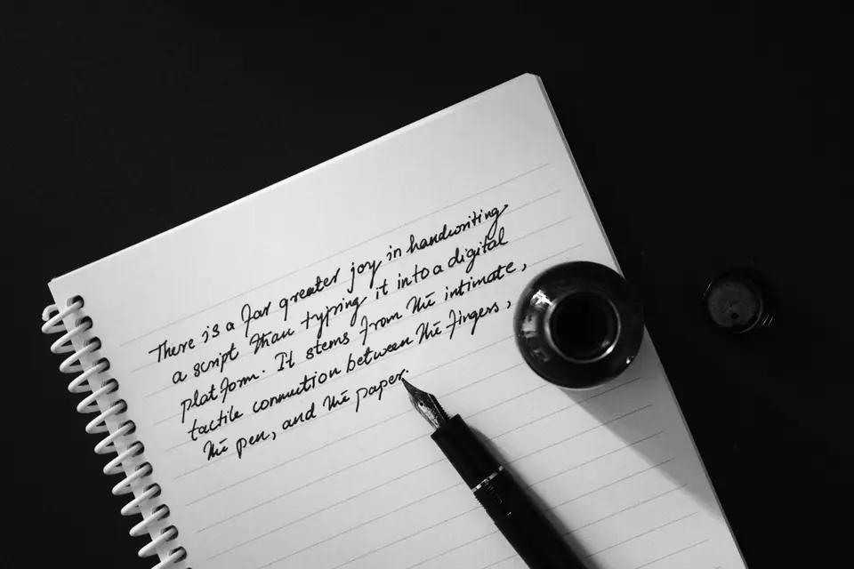 A fountain pen rests on a spiral notebook with handwritten lines beside a small inkwell on a dark surface.