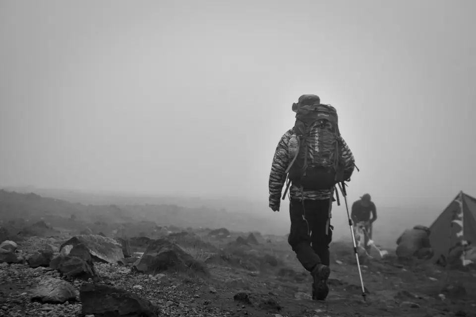 A hiker with a large backpack and trekking pole walks across rocky ground in fog.