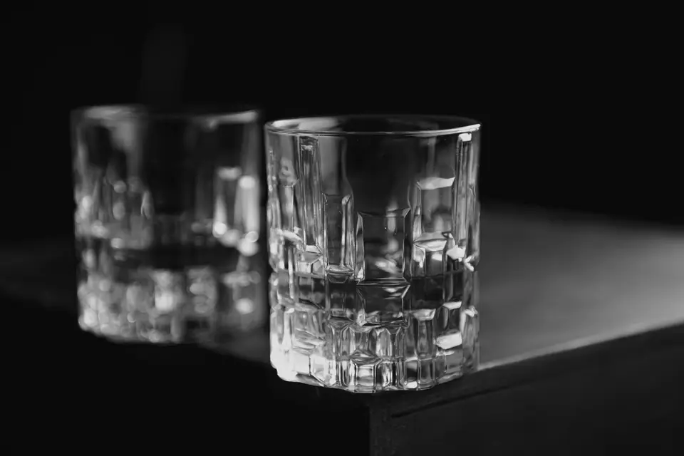 Two clear, heavy-bottomed glass tumblers sit on the corner of a dark table against a black background.