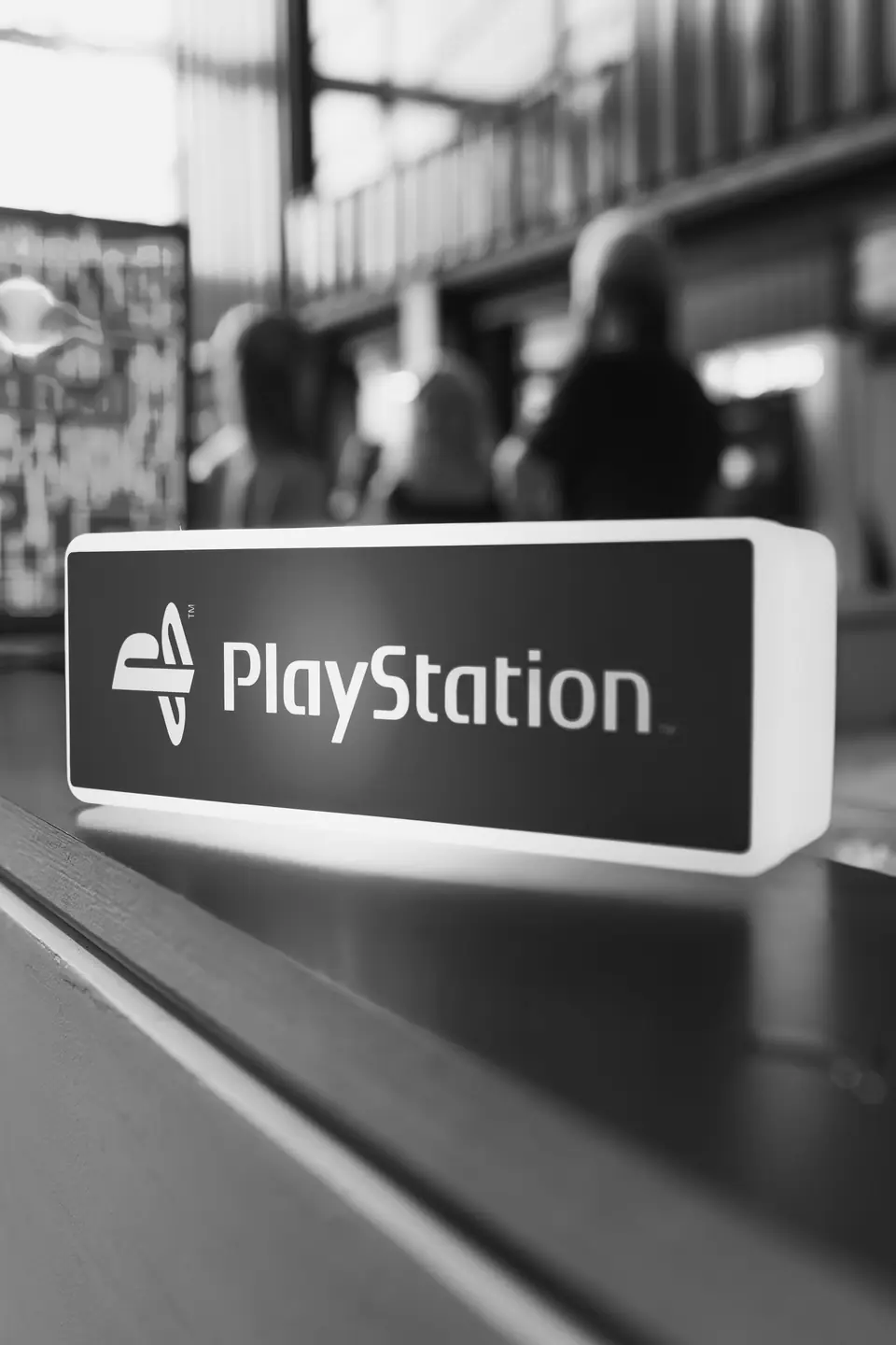 A lit rectangular sign reading “PlayStation” sits on a counter with blurred people behind it.