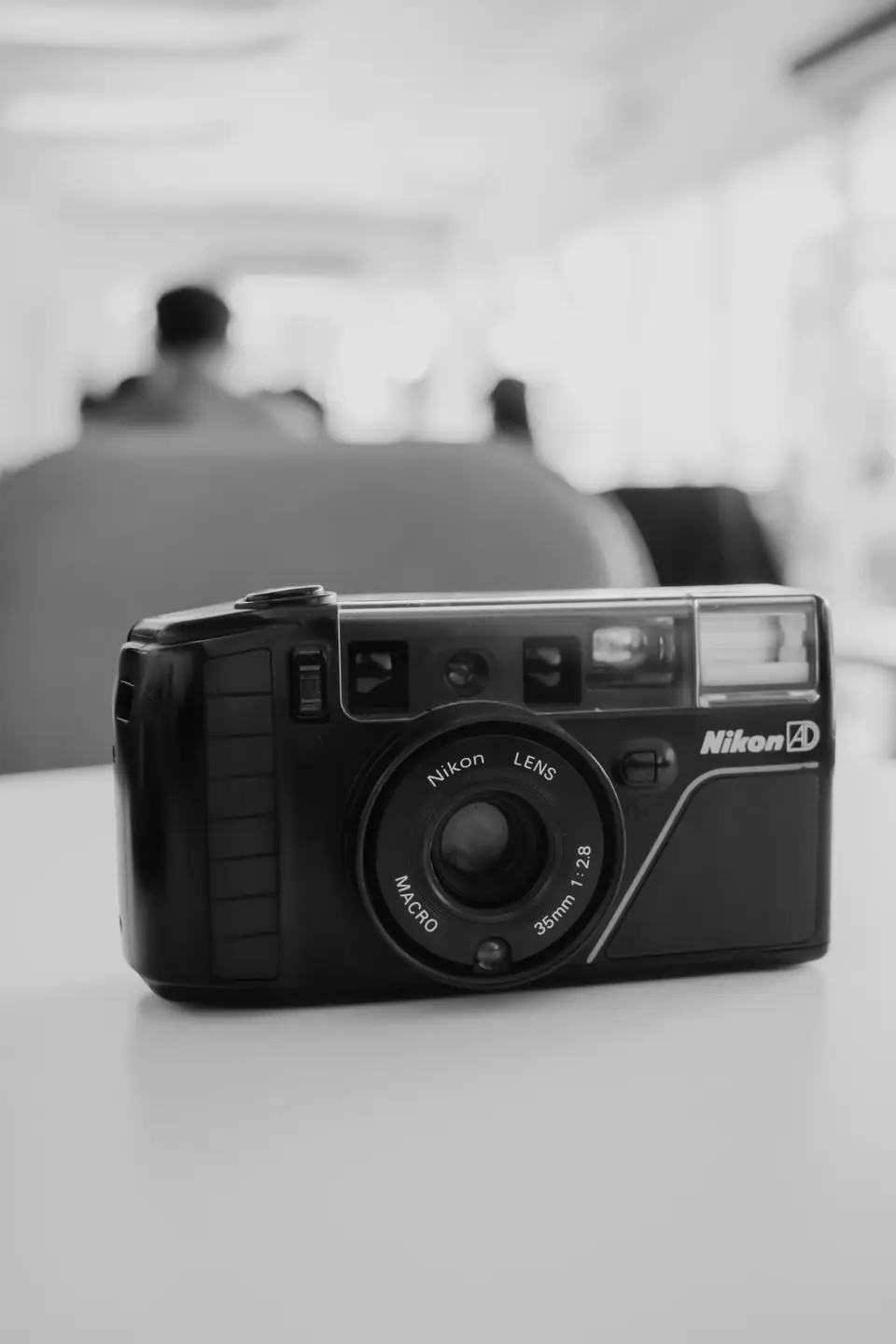 A black Nikon compact film camera sits on a white table with a blurred room behind it.