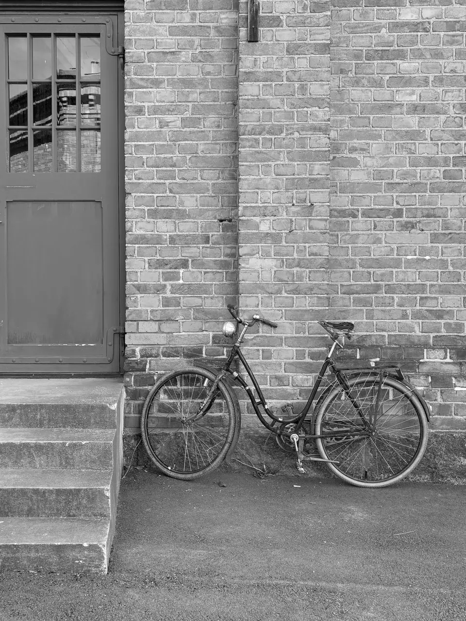 An old step-through bicycle leans against a brick wall next to a doorway and concrete steps.