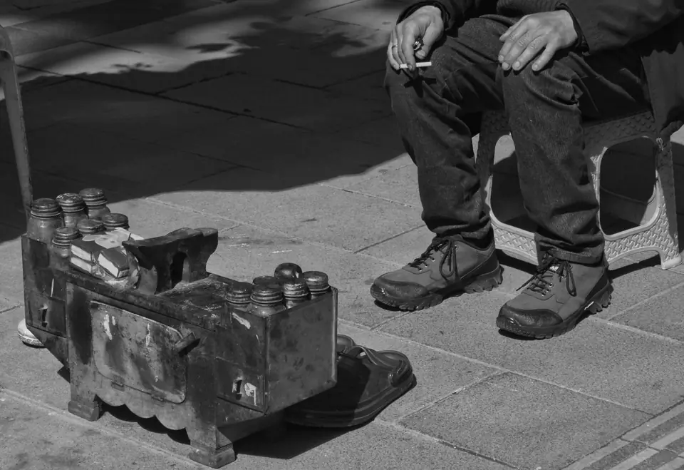 A wooden shoeshine box with tins sits on a paved sidewalk beside a seated man in sneakers holding a cigarette.