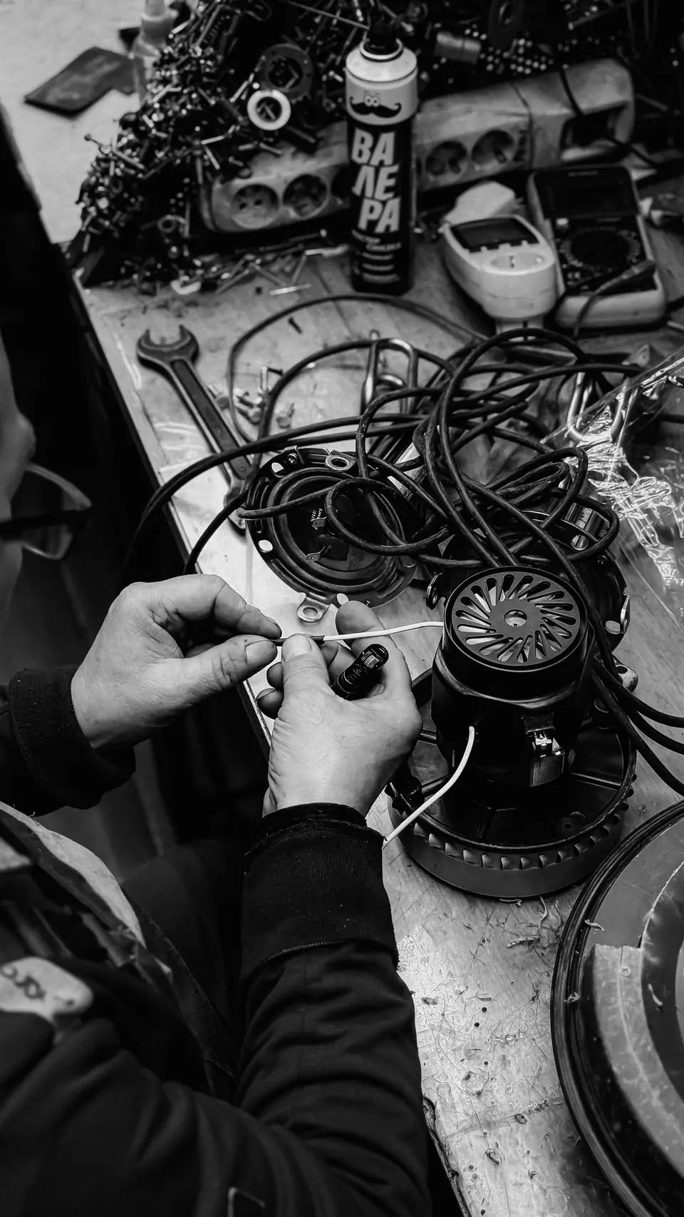 Hands work on wires and a small motor on a cluttered workbench covered with tools.