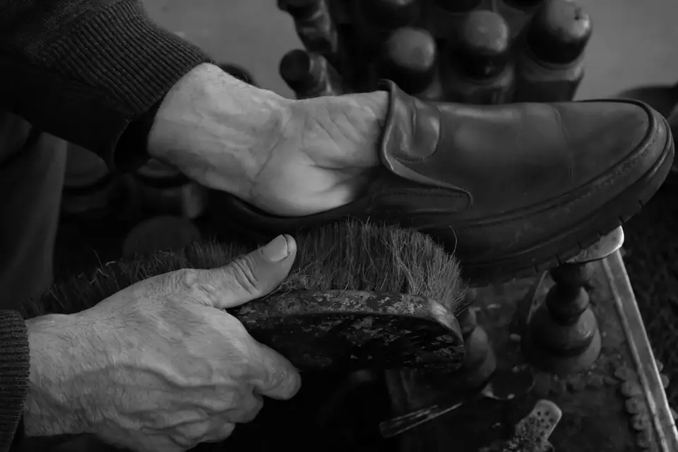 A man’s hands buff a leather loafer with a wooden-backed brush on a shoeshine stand with tins behind.