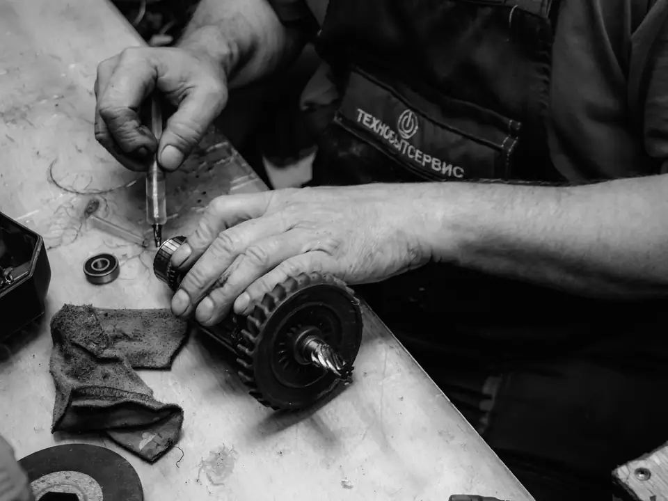 A worker’s hands use a screwdriver on an electric motor armature at a grimy workbench with a rag.