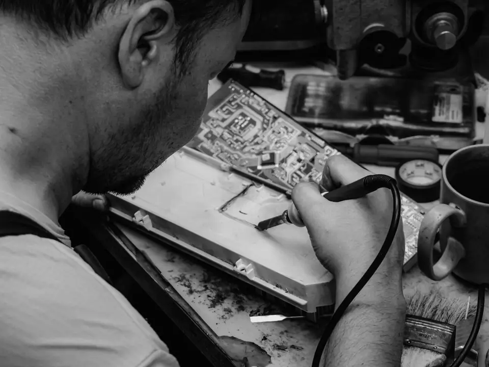 A man leans over a workbench, soldering a green circuit board with a hand iron.
