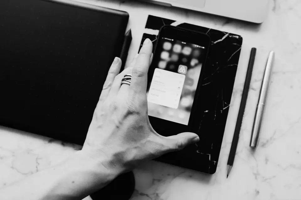 A hand with a ring touches a smartphone showing open apps, next to a notebook, pencil and pen.