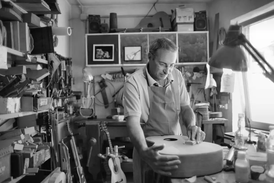 A man in an apron works on a wooden guitar body at a bench in a busy instrument workshop.