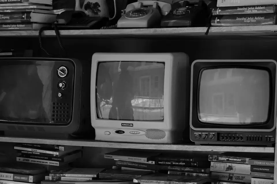 Three small vintage television sets sit on a shelf between rows of books and old rotary telephones.